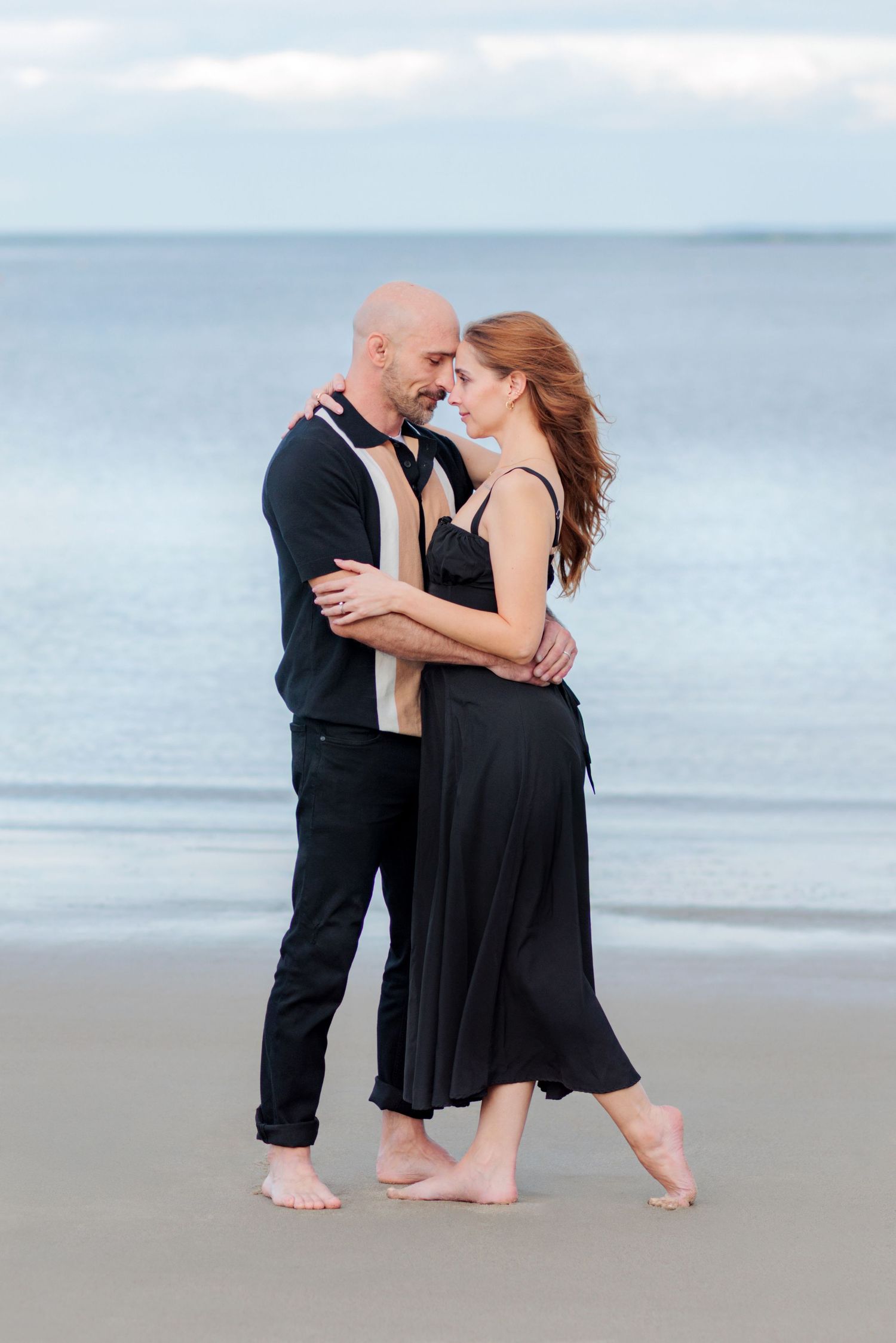 A couple in black attire shares an intimate dance moment barefoot on a misty beach at dusk.