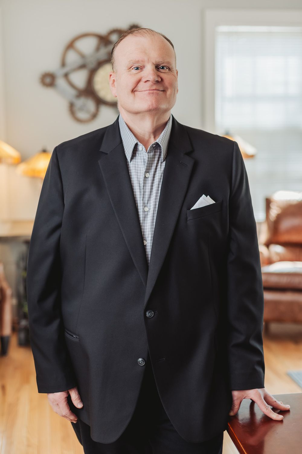 Smiling middle-aged man in black blazer with white pocket square standing in a well-lit home interior.