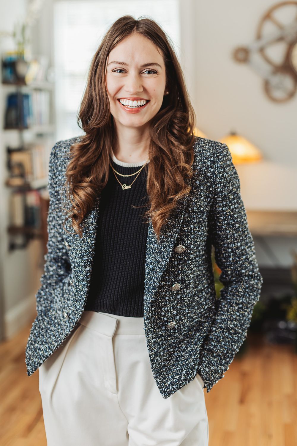 Smiling woman in a tweed blazer and white pants stands confidently in a bright, stylish office space.