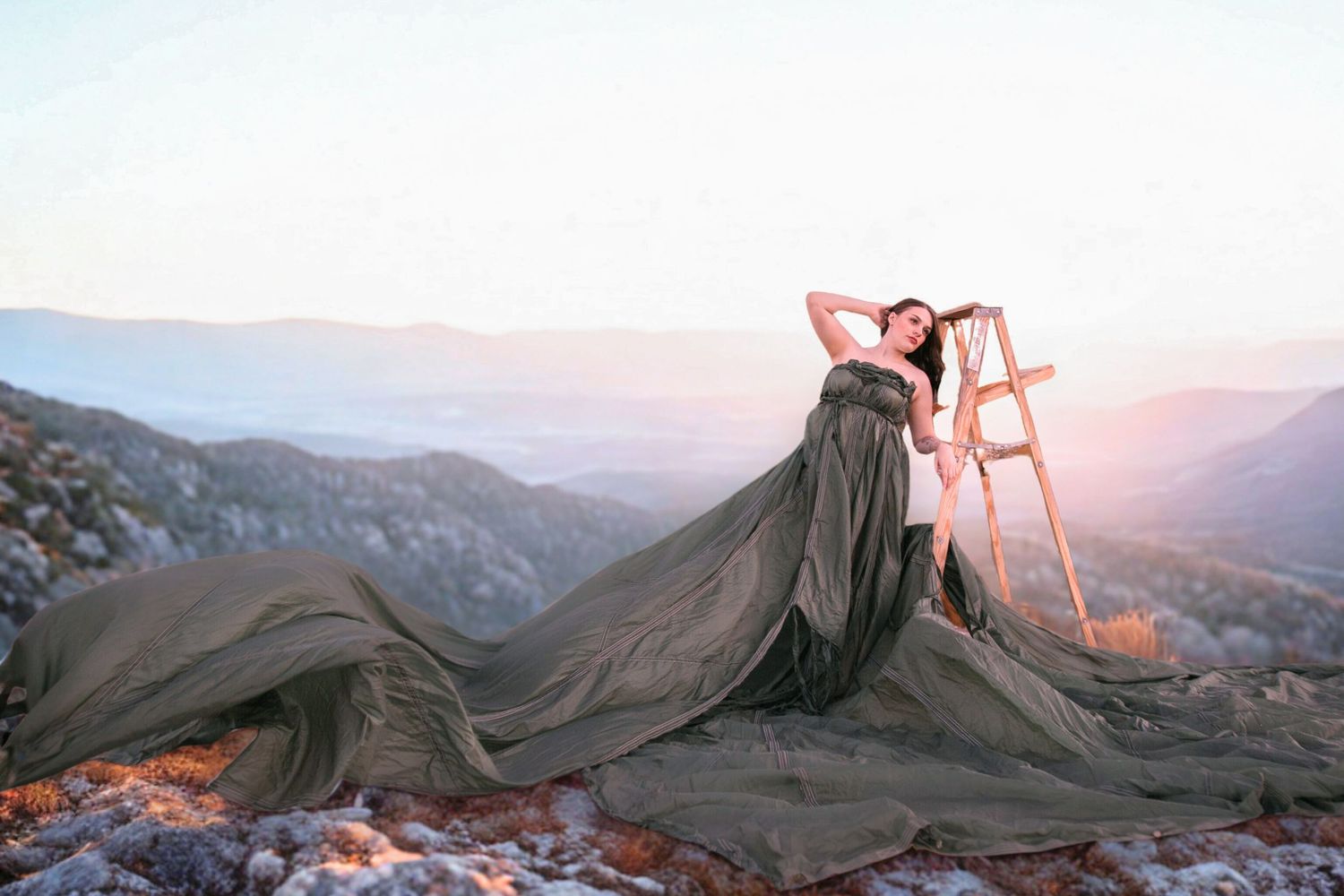 Woman in flowing olive green gown posing dramatically on rocky mountain summit at sunset.