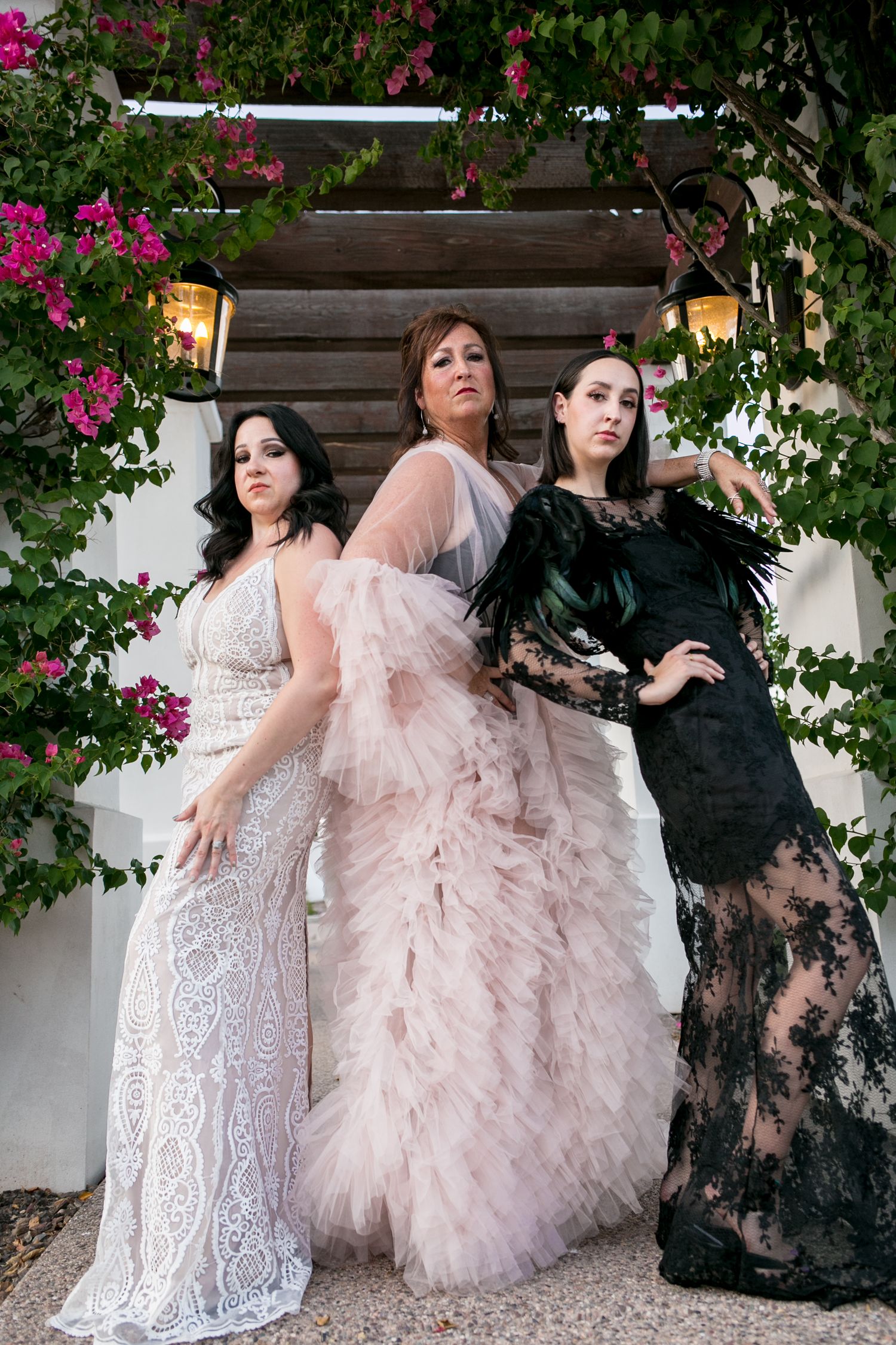 Three women in elegant white lace, pink tulle, and black lace gowns under floral arch.