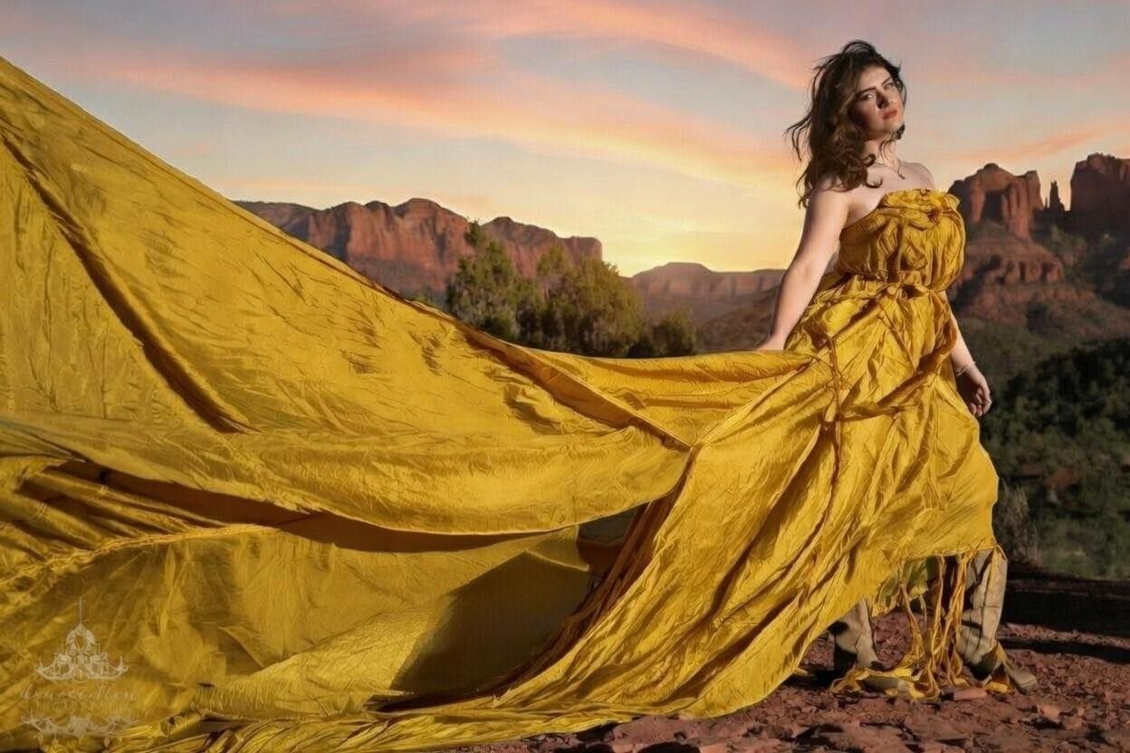 Woman in golden yellow billowing dress poses dramatically against red rock canyon at sunset.