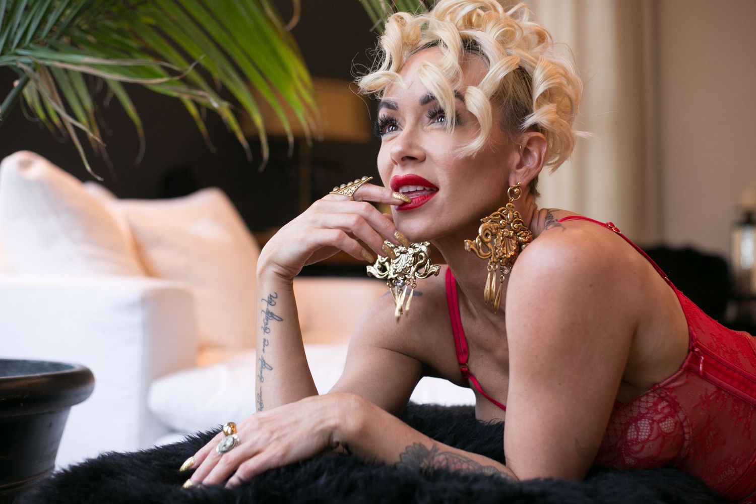 Glamorous woman in red lace lingerie with gold earrings and blonde updo lying on dark fur surface.