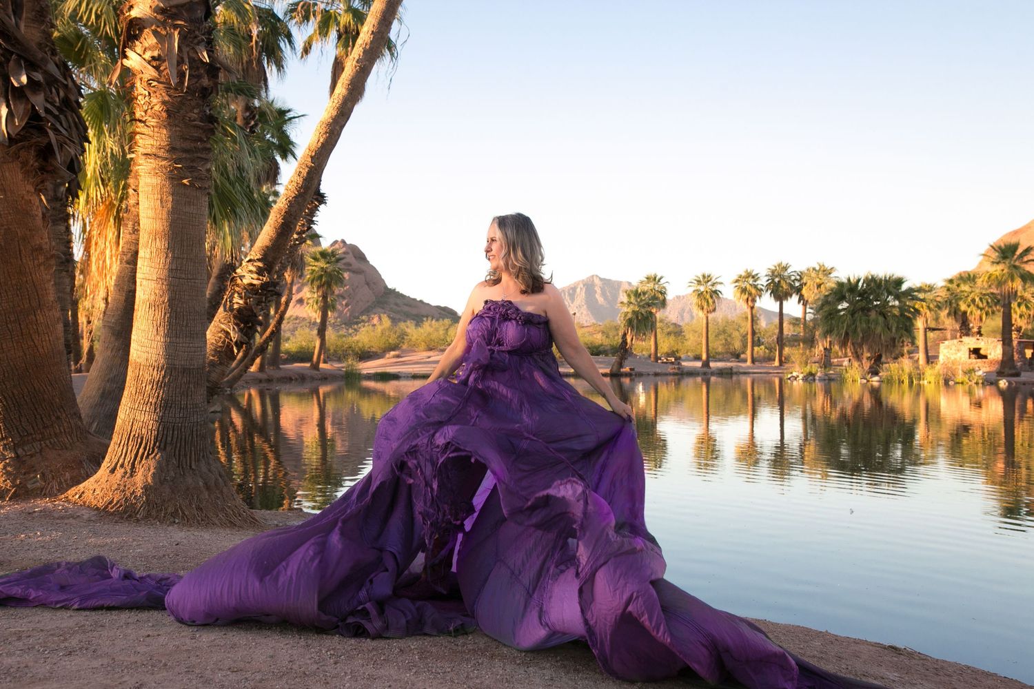 Woman in stunning purple ball gown sits by reflective lake surrounded by palm trees and mountain backdrop.