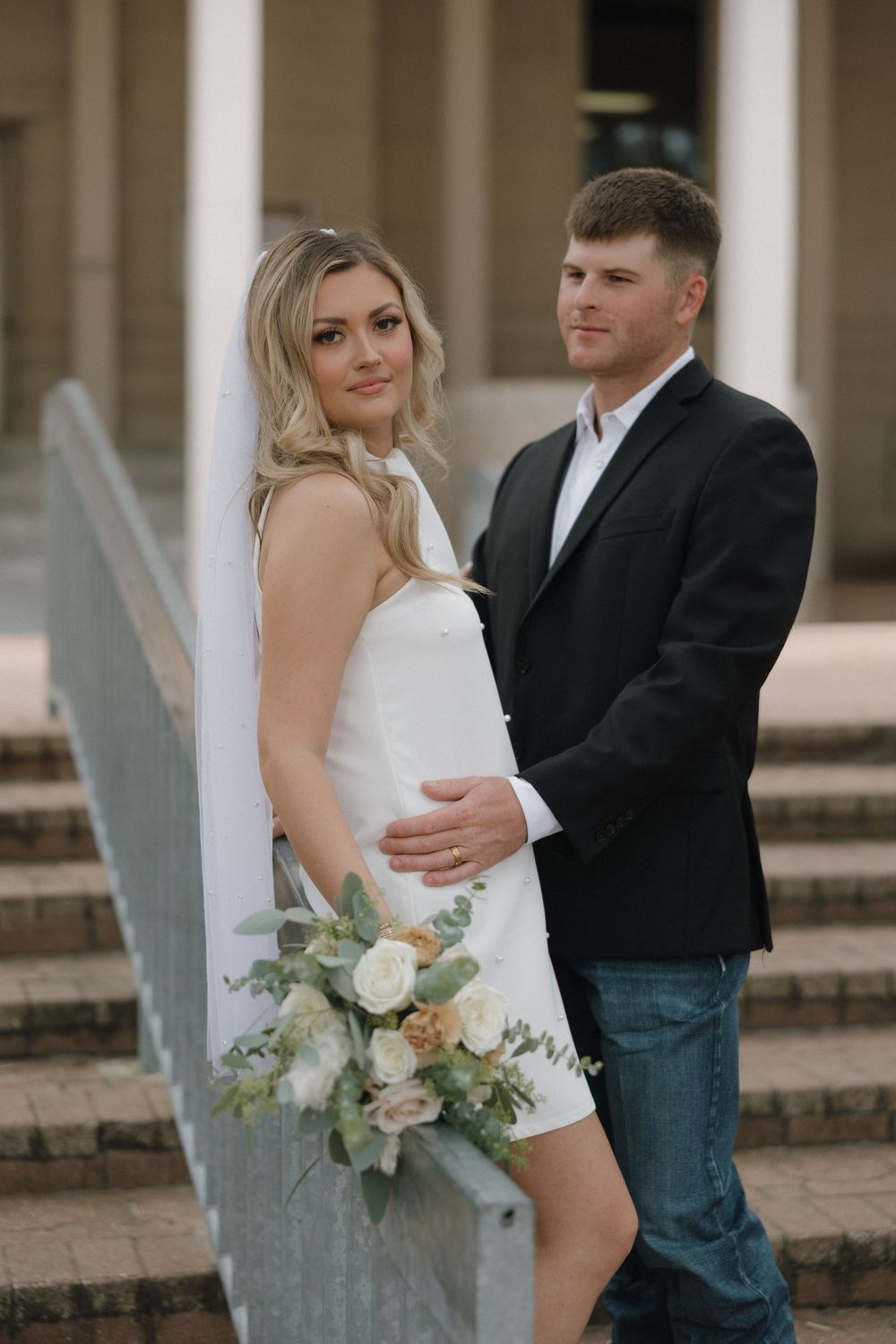 A couple in wedding attire poses on steps of a courthouse, with the bride holding a pastel floral bouquet.