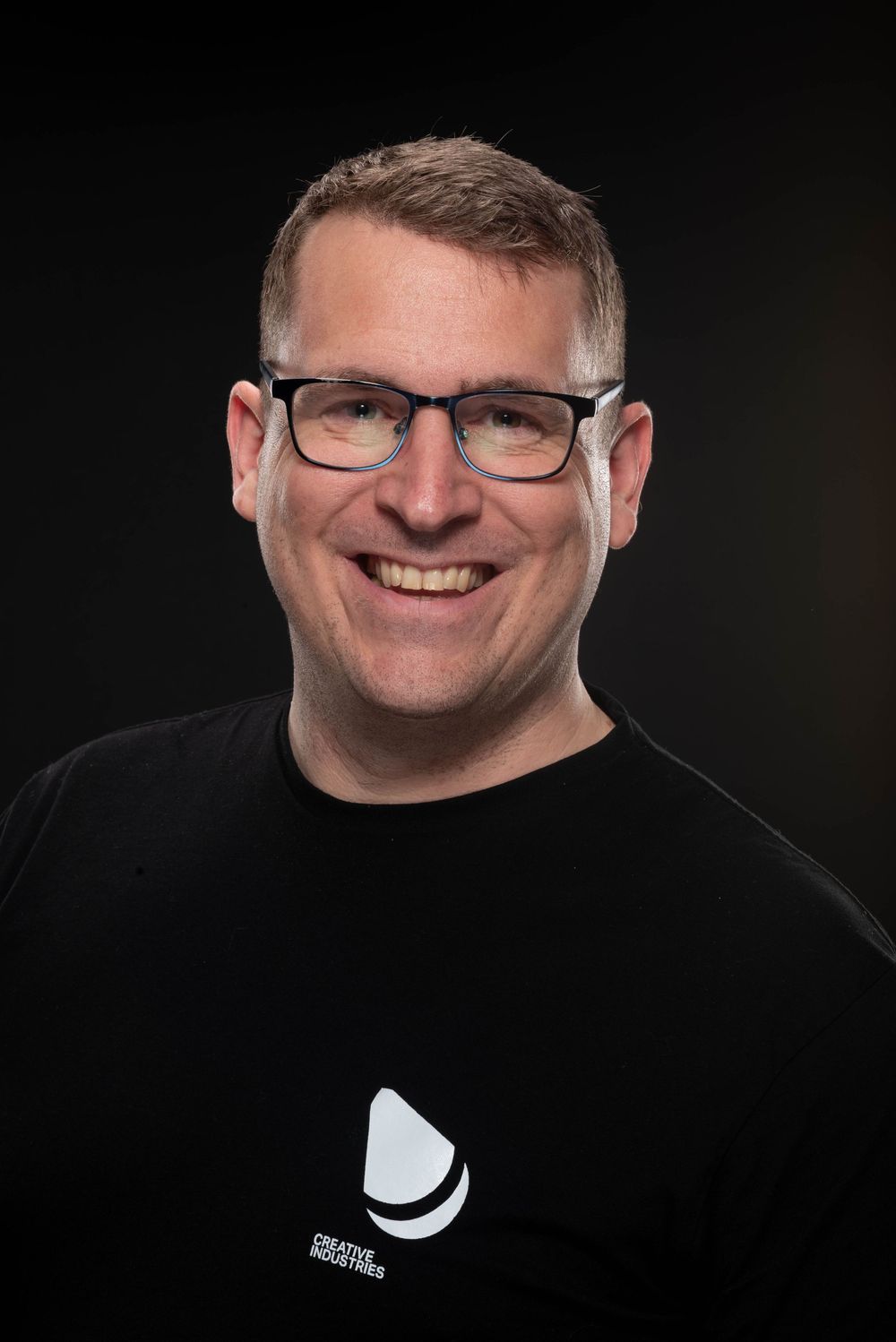 Professional headshot portrait against black background showing person wearing glasses with warm smile.