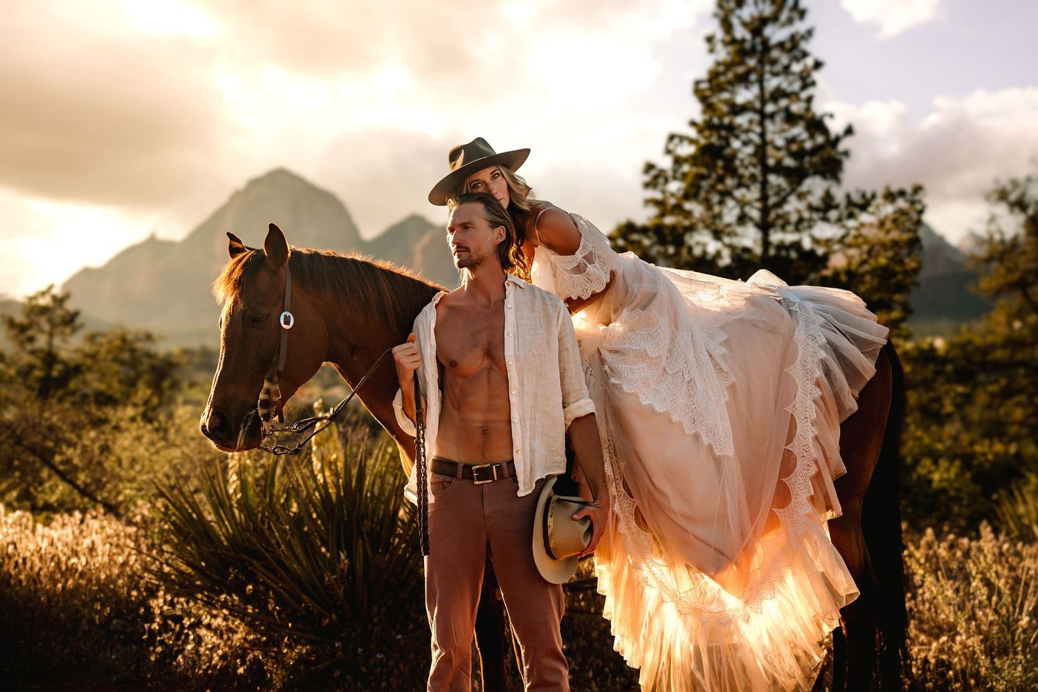 Shirtless cowboy stands with brown horse at golden sunset in desert mountains, wearing hat and holding bridal gown.