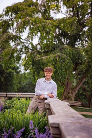 Young man in white shirt leaning on wooden bridge railing surrounded by purple flowers and lush green trees.