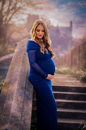 Pregnant woman in blue off-shoulder gown cradling baby bump on stone steps at golden hour.