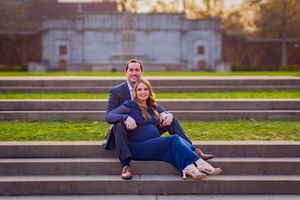 Couple posing on outdoor stone steps, woman in blue jeans leaning against man in navy suit, green lawn background.