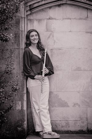 Black and white photo of a smiling young woman holding a flute, leaning against a stone wall outdoors.