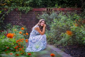 Woman in floral dress sitting in a lush garden surrounded by orange marigolds and green foliage near a brick wall.