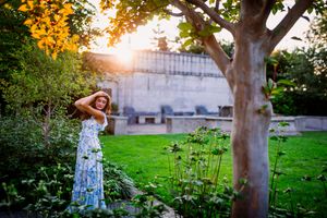 Woman in floral maxi dress and hat poses in lush garden at golden hour with sunlight streaming through trees.