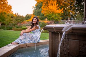 Young woman in floral dress sitting by a stone fountain in a lush garden with autumn foliage at sunset.