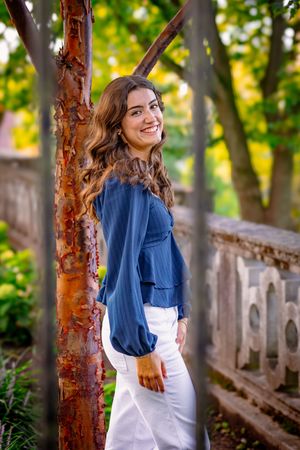 Young woman in blue top and white pants smiling outdoors near rustic tree and stone railing.