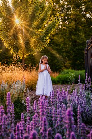 Girl in white dress praying among purple lavender flowers at golden sunset in a lush garden.