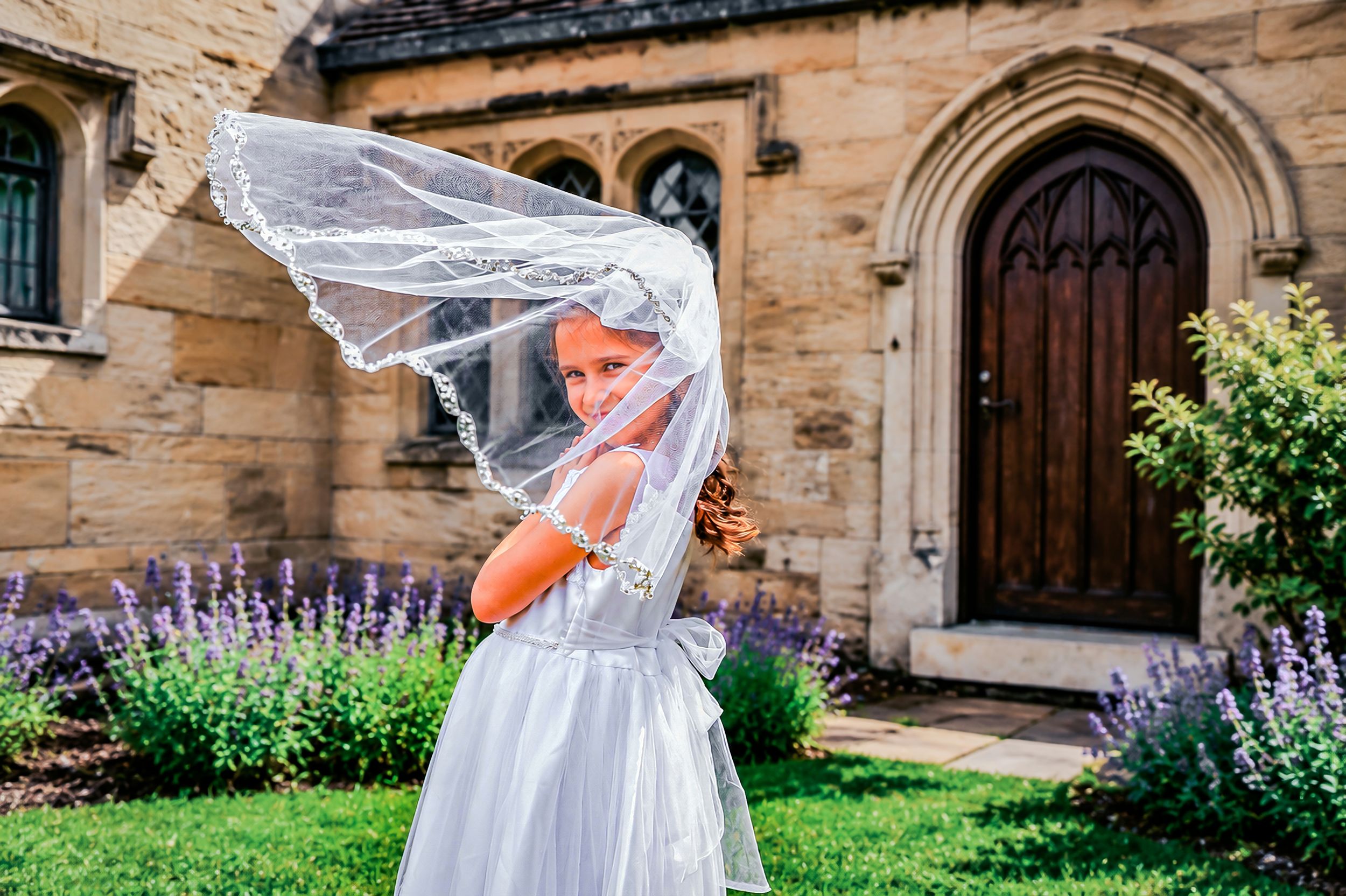 Young girl in white dress and veil stands outside stone church with purple flowers, veil blowing in wind.