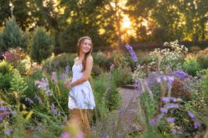 Young woman in white dress smiling in a lush garden with purple wildflowers at golden hour sunset.