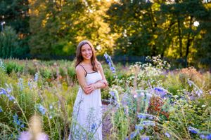 Young woman in white dress smiling among wildflowers in a sunlit garden at golden hour.