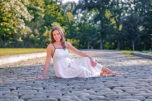 Young woman in white dress sitting on cobblestone path in sunny park, smiling cheerfully at camera.