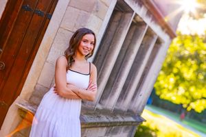 Young woman in white dress poses with arms crossed beside stone columns in golden sunlight.