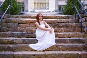 Young woman in white dress sitting gracefully on stone steps outdoors, smiling at camera during senior portrait session.