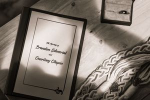 Black and white wedding program for Brandon Schmersal and Courtney Chapin beside a ring box on a wooden table.