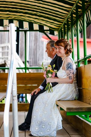 Bride in white floral dress holding bouquet sits with older man in tuxedo on green striped bench at outdoor wedding.