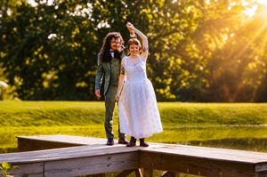 Newlywed couple dancing on wooden bridge at sunset, bride in white dress, groom in green suit, lush green park background.