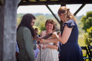 Three women share a joyful moment during an outdoor wedding ceremony, exchanging rings under a rustic wooden structure.