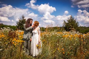 Bride and groom embracing in a vibrant wildflower meadow under a blue sky with fluffy white clouds.