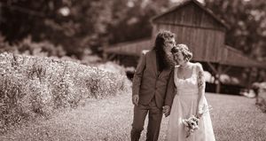 Couple holding hands in wedding attire walking near rustic barn surrounded by lush greenery in sepia-toned photo.
