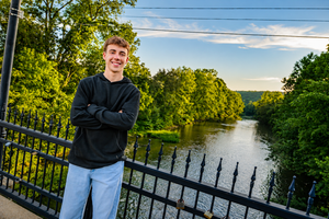 Young man in black hoodie stands arms crossed on bridge overlooking scenic river surrounded by lush green trees.