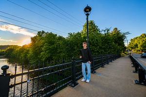 Man standing on a scenic riverside walkway with iron railings, lamp post, and lush green trees at sunset.