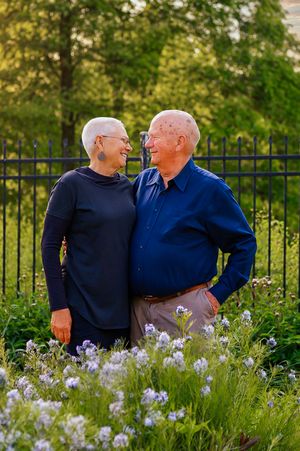 Elderly couple smiling at each other in a garden with purple wildflowers and green trees in the background.
