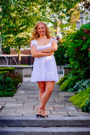 Young woman in white mini dress with arms crossed, smiling in a lush green garden pathway.