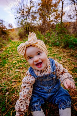 Cheerful baby girl in denim overalls and yellow bow headband sitting outdoors in autumn nature setting.