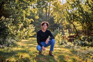 Young person with curly hair smiling, crouching in a sunny green garden wearing jeans and a black top.