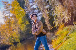 Young person smiling outdoors in autumn park, wearing brown jacket and jeans, with colorful fall foliage and river behind.