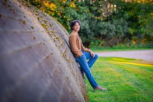 Young man in brown jacket and jeans leaning against a curved stone wall in a lush green park setting.
