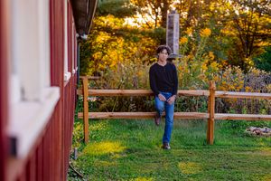 A young man in jeans and black sweater leans on a wooden fence near a red barn at golden hour.