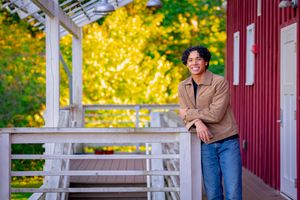 Young man in brown jacket leaning on white wooden deck railing with red barn and autumn trees behind.