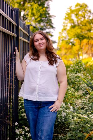 Young woman in white blouse and jeans leaning against black iron fence surrounded by autumn foliage.