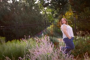 Woman in white top and jeans poses among purple wildflowers in a sunlit garden with iron fence.