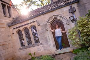 Woman posing in doorway of historic stone building with arched windows and warm sunset light filtering through trees.
