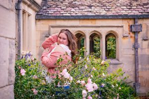 Woman in pink coat smelling roses in a cottage garden with stone building and arched windows backdrop.
