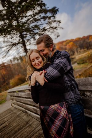 Couple embracing warmly outdoors in autumn, woman smiling with eyes closed, colorful fall foliage in background.