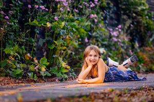 A smiling young girl in a floral dress lying on a garden path surrounded by lush green plants and pink flowers.