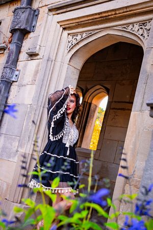 Woman in black vintage dress poses gracefully beneath ornate stone arch of historic castle with blue flowers in foreground.