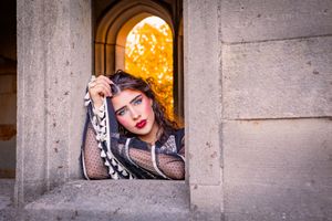 Woman in mesh top with bold makeup poses in stone archway with autumn foliage backdrop.